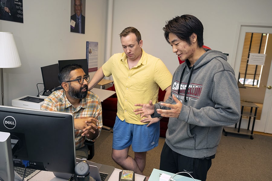 Bo Xie stands next to his supervisors Max Holmes and Ani Kapuria, in front of Ani’s desk. They are engaging in a conversation.