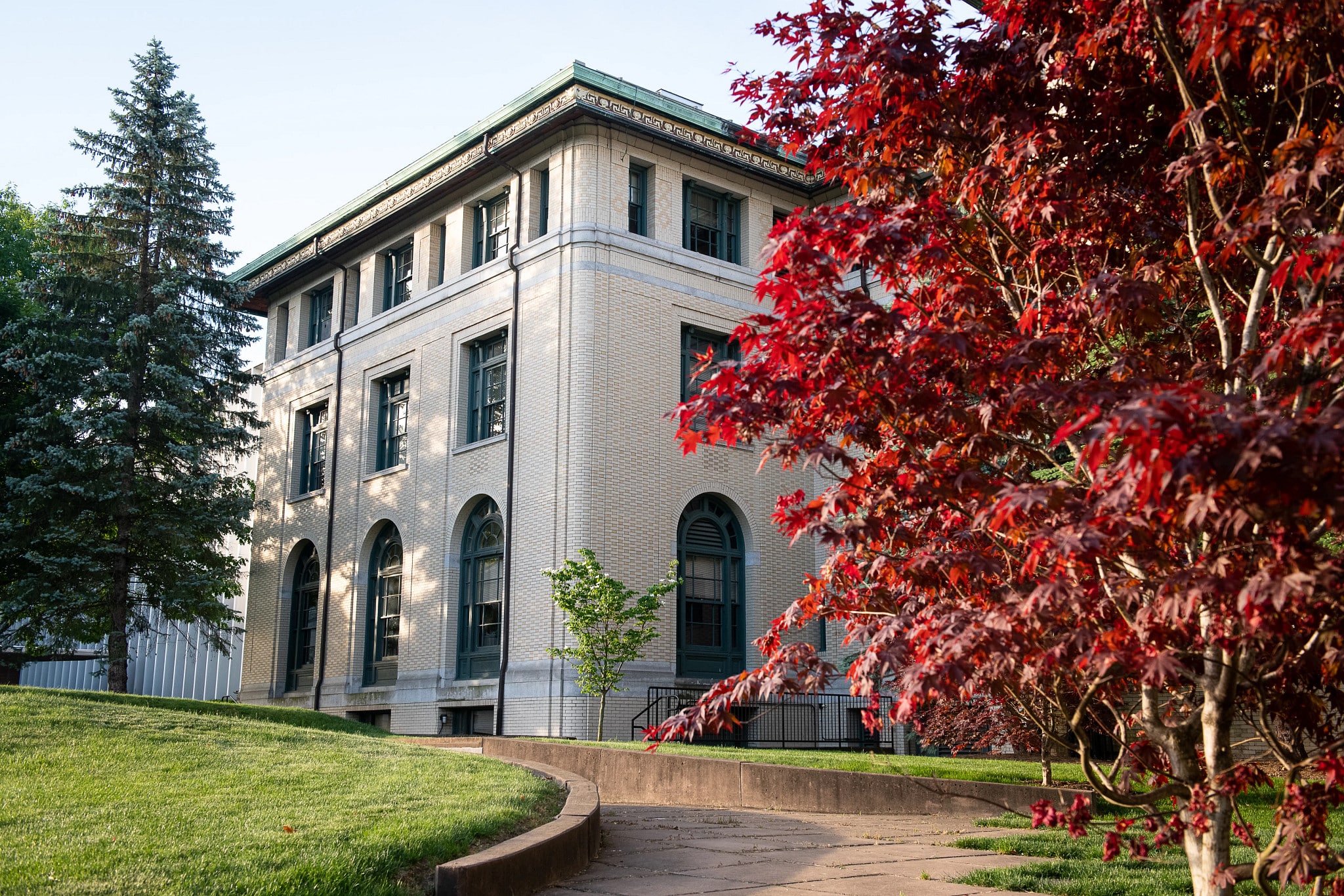 A brick building on campus with a green roof and white trim stands on a grassy slope. Several trees with red leaves are in the foreground.