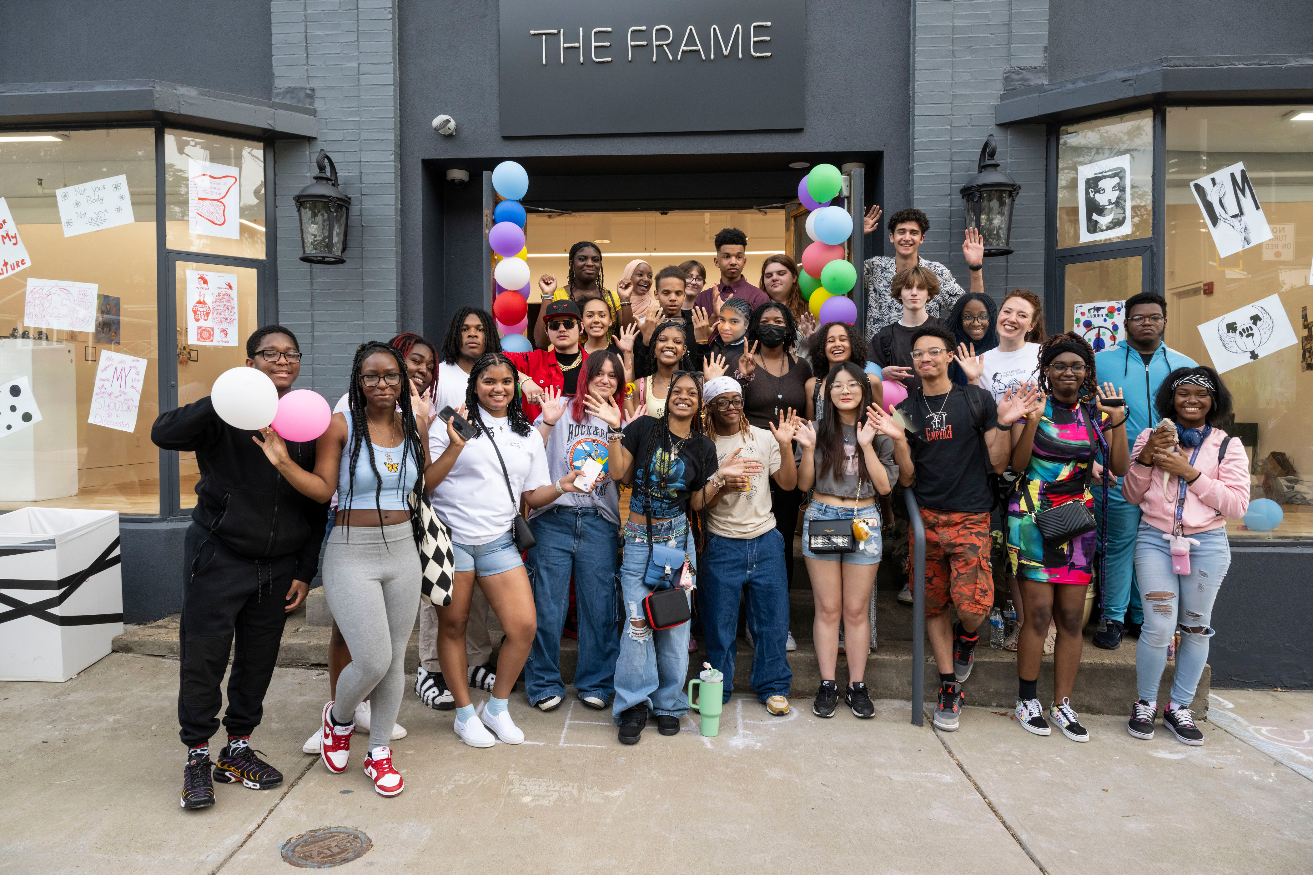 LEAP students and their teacher posing together outside of The Frame Gallery during their Showcase celebration. There are balloons around the door and artwork in the windows.