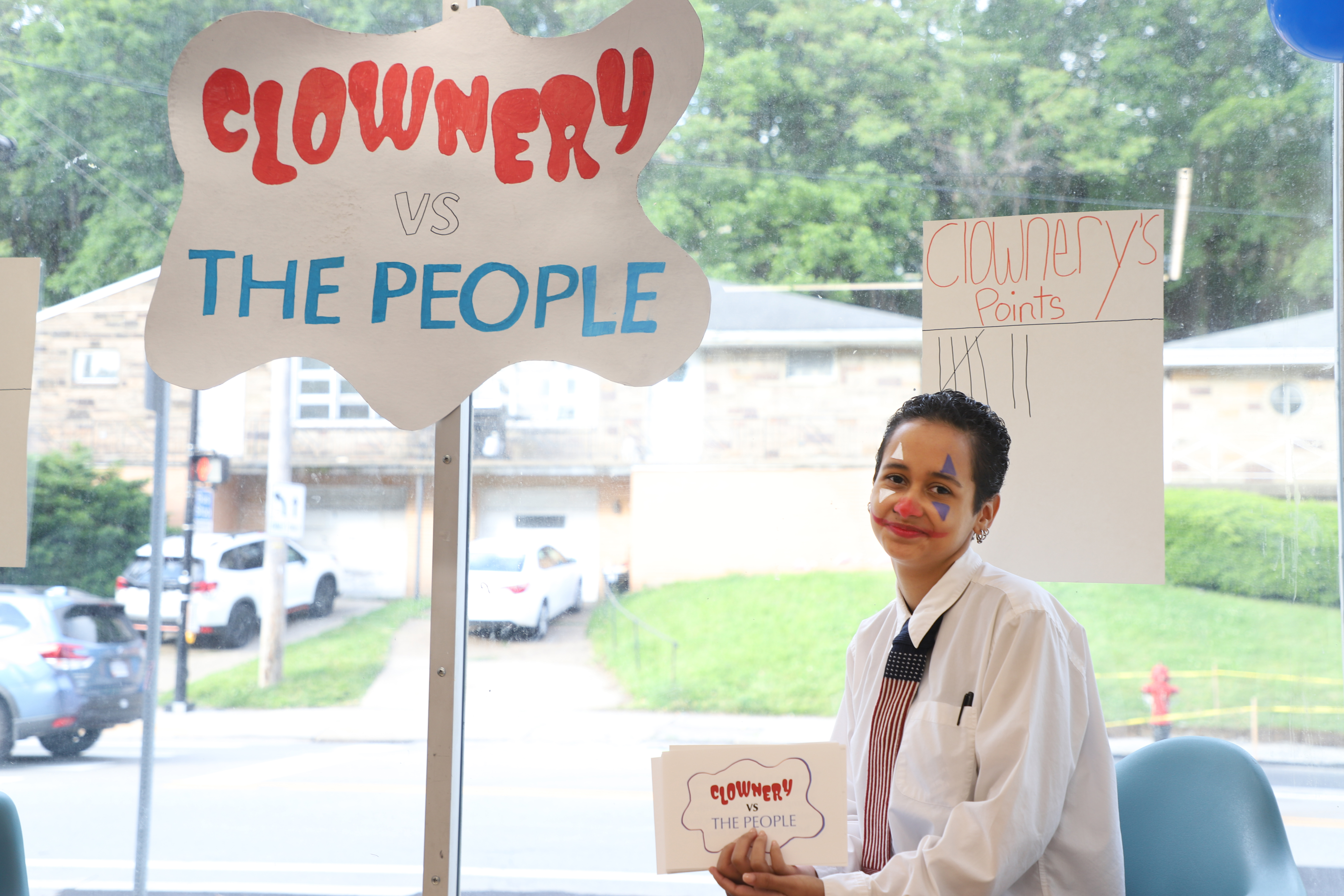 LEAP student Alex wearing clown makeup and sitting in front of a wavy sign that reads "Clownery vs. The People"