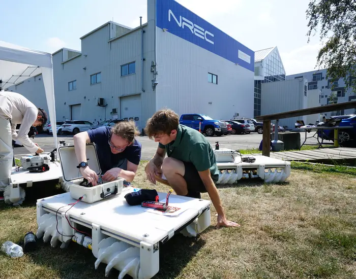 Two students assemble a robot in front of a building labeled NREC