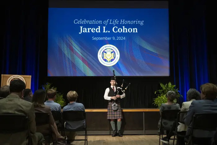 A man stands in front of a stage playing bagpipes dressed in traditional Scottish garb, including a kilt. Behind him, a screen displays a slide that says, "Celebration of Life Honoring Jared L. Cohan - September 9, 2024"