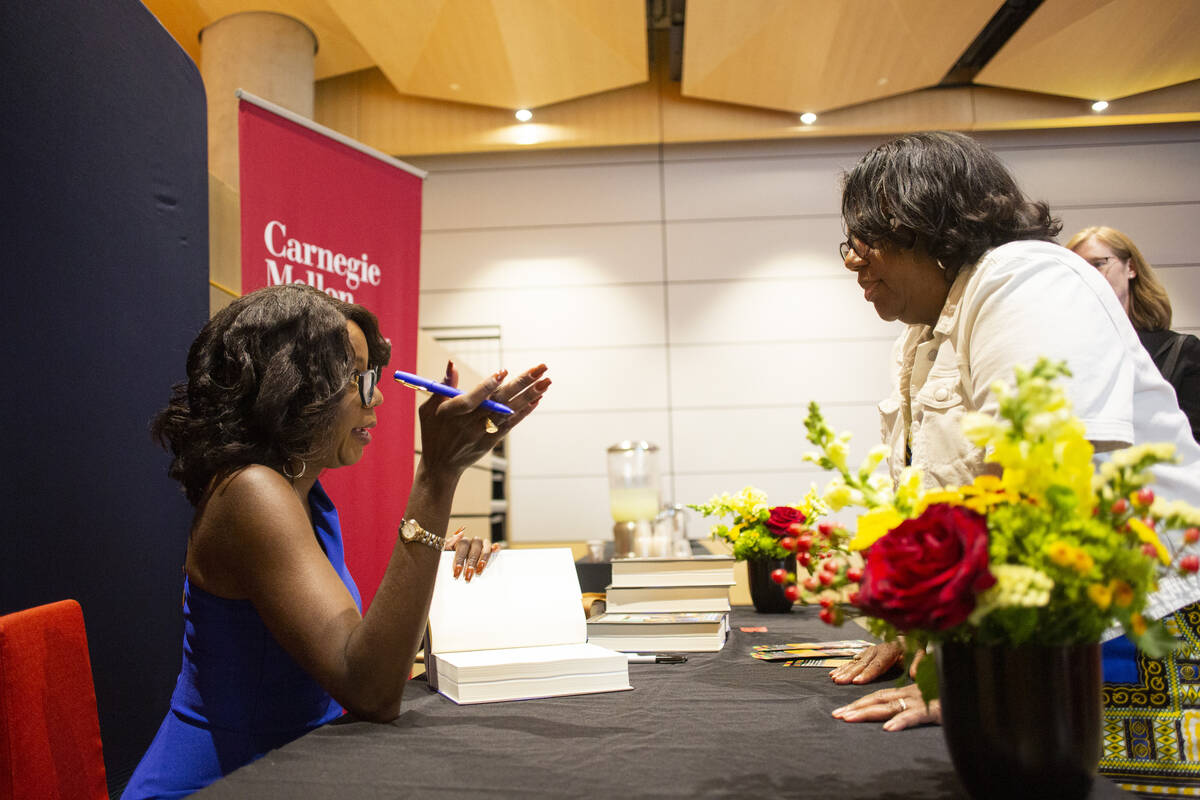A Black woman with softly curled hair sits behind a table and gestures with her elbow on the table and a pen in her hand while holding open a book with the other. She talks to a Black woman standing behind the other side of the table, which has a stack of books and a small vase of red and white flowers.