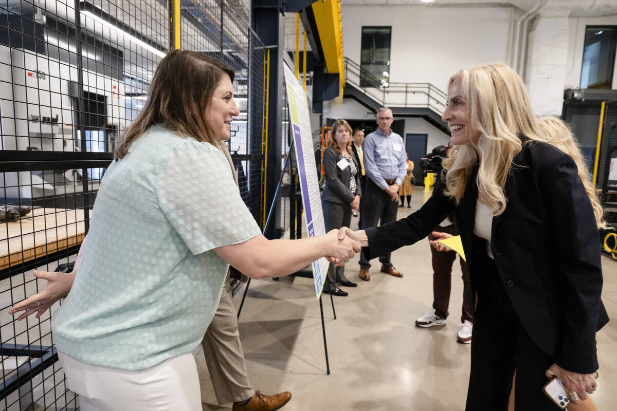 A white woman with brown hair in a light green top and white pants shakes hands with a white woman with wavy blonde hair wearing a black suit, seen from the side in an industrial setting with people behind them looking on.