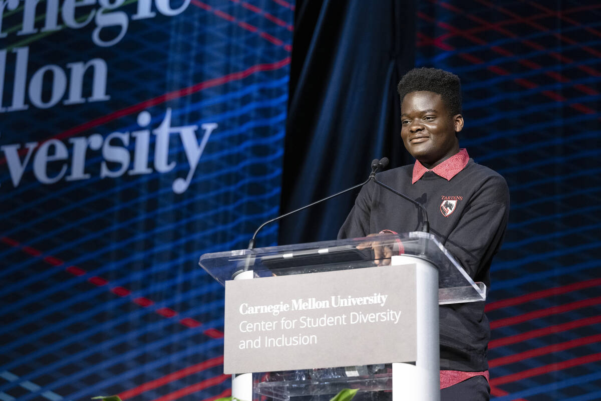 A Black man wearing a black sweatshirt with red collar stands behind a clear acrylic podium with a sign that says "Center for Student Diversity and Inclusion"