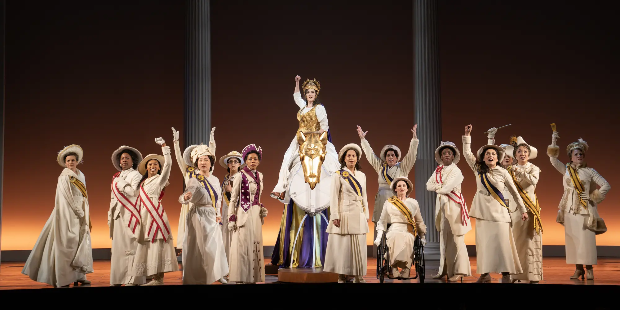 About 15 women, all dressed in ivory wearing hats and sashes to emulate the 1920s, stand on a stage, looking toward the audience. One woman in the center, on top of a prop made to look like a horse, wears a gold headdress and top akin to a Greek goddess.