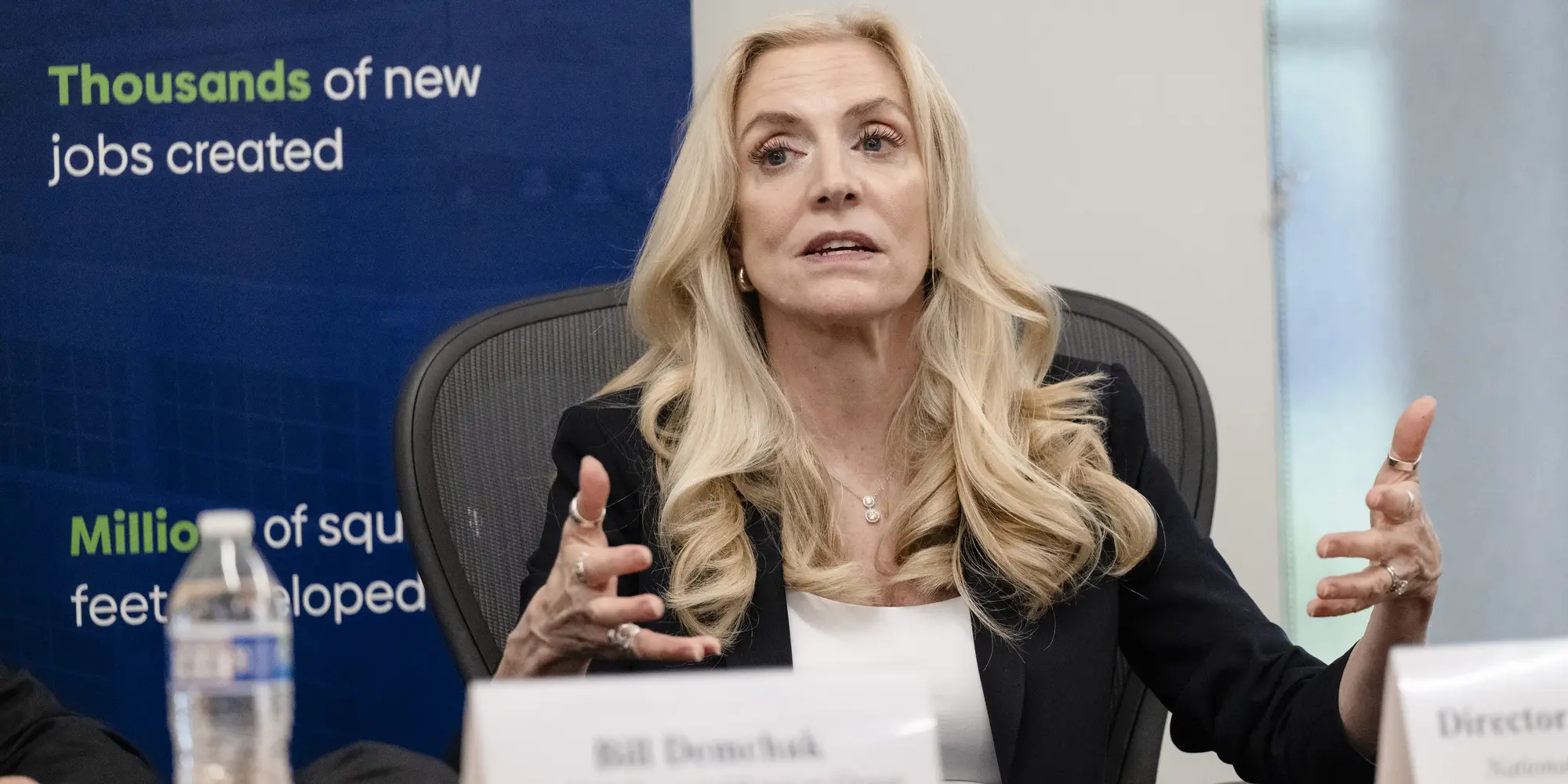 A white woman with long wavy blonde hair sits at a table behind a name placard with her hands raised to gesture while speaking.