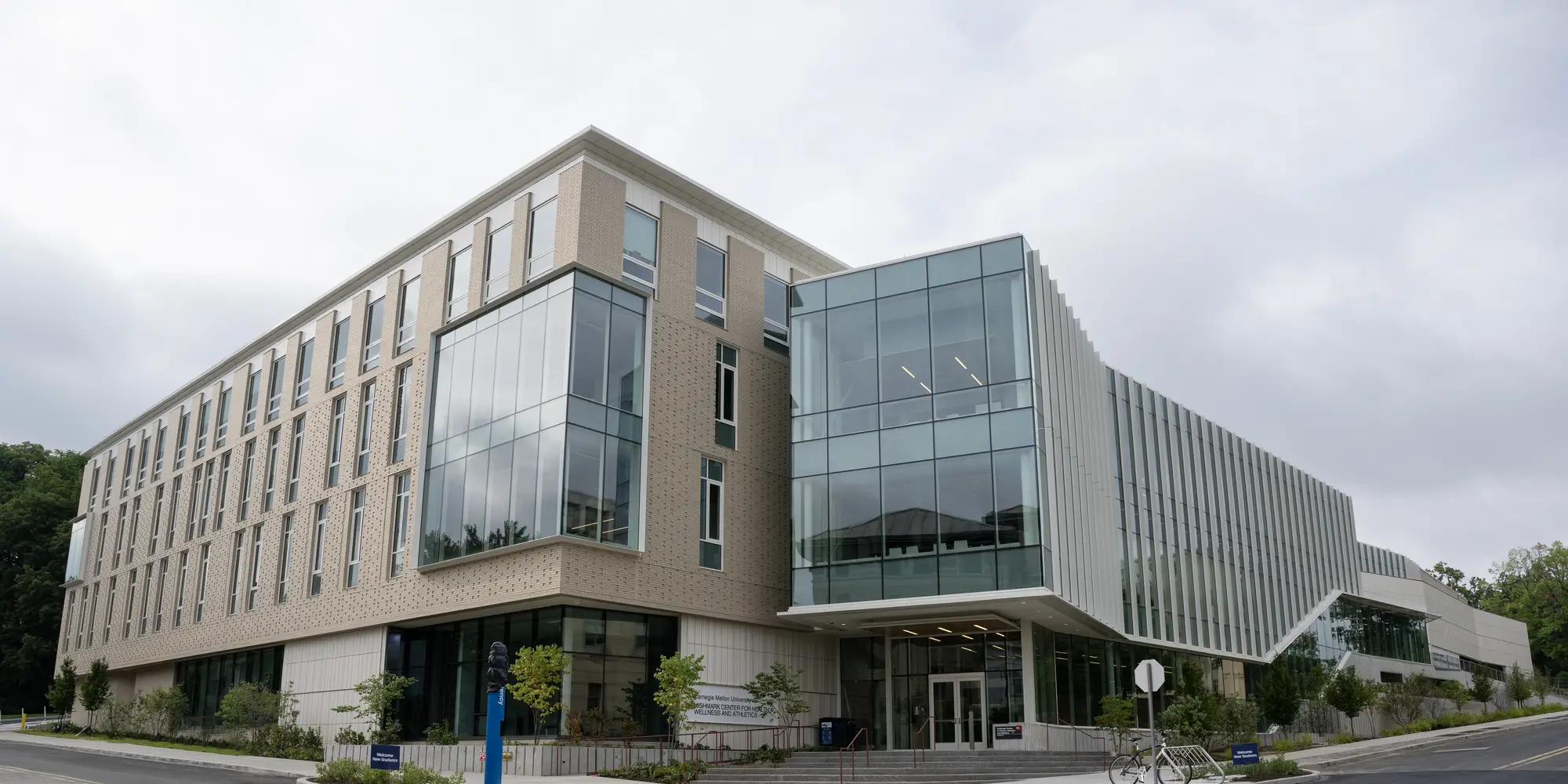 The exterior of a four-story building from a streetview of the intersection of roads where it sits, with a pattern of beige-colored brick covering the left side while a protruding cube of glass windows encompasses most of the right side.