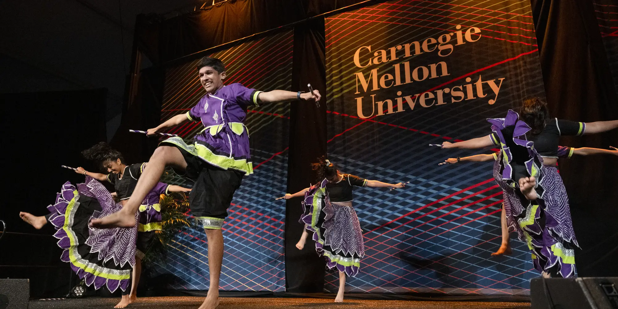 Five people in purple and green costumes kick their foot together as part of a dance group on a stage with a backdrop behind them that says 