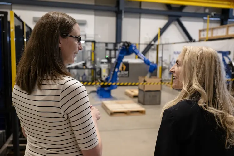 A white woman with shoulder-length brown hair, a white and gray striped shirt and glasses talks with a white woman with long blonde hair in a black suit jacket, while in between them in the background stands a large blue robotic arm with boxes and a wooden pallet nearby.