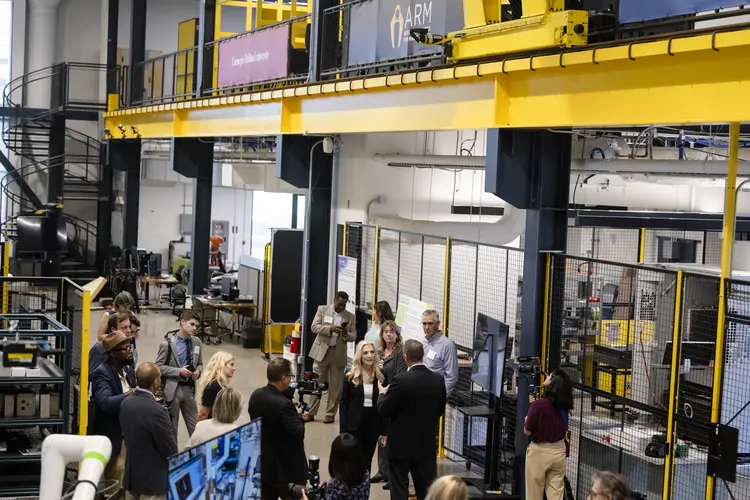 A group of about 20 people stand and talk inside an industrial-looking space with black wire area dividers and several computer screens, while a steel beam painted yellow frames the top of the image.