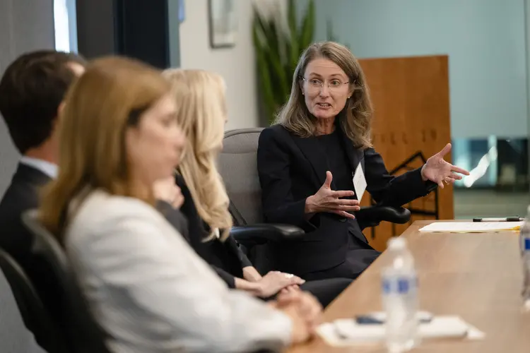 A white woman with dark blonde hair gestures in the background while three people next to her out of focus in the foreground listen.