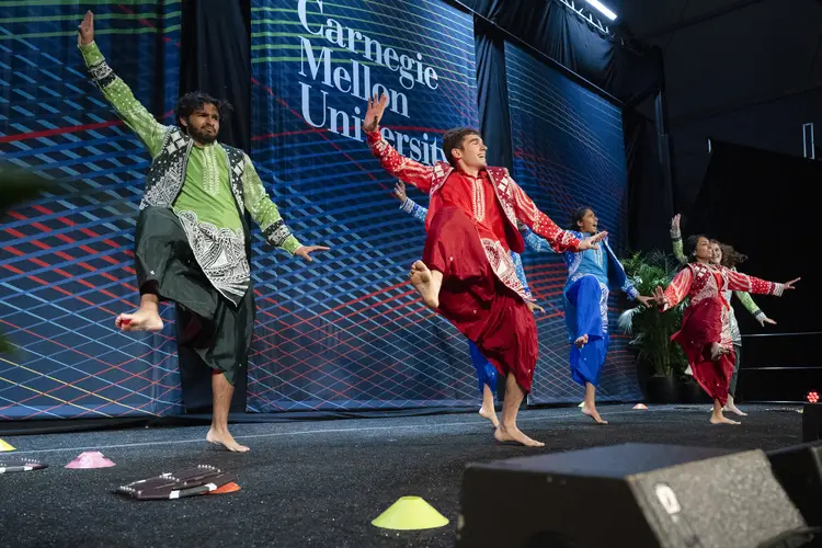 Six people, each dressed in a different brightly colored costume, dance with their left knee raised to their chest and foot raised to knee height.