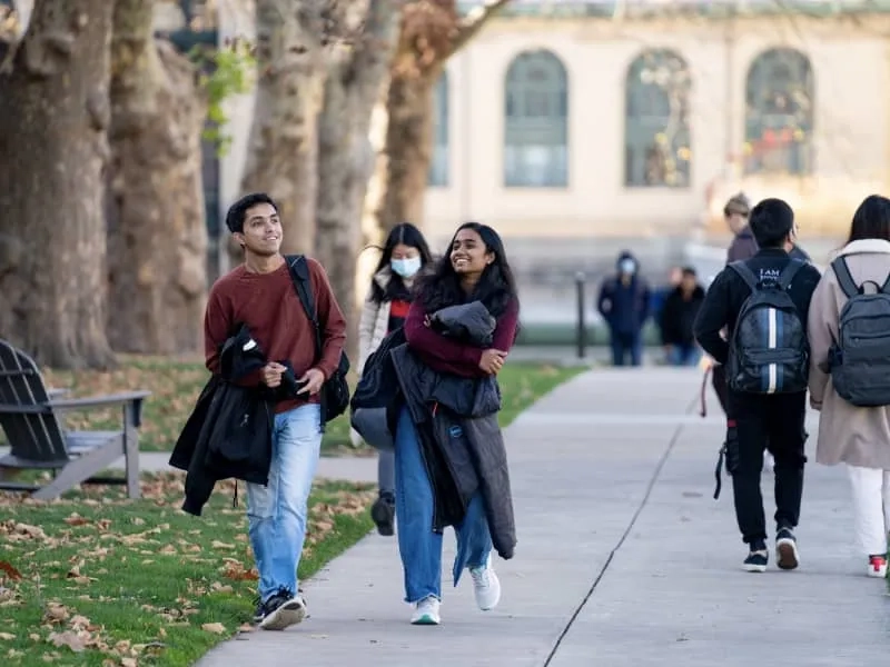 Two students smiling and wearing backpacks while walking along a pathway on CMU's campus with additional students in the background