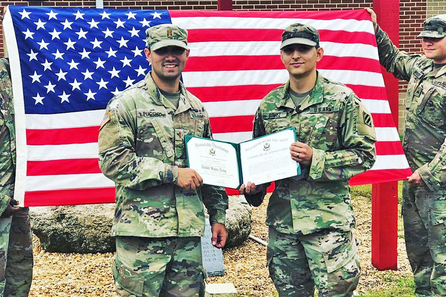 travis in camouflage uniform with military colleague holding diploma in front of the United States flag smiling at camera